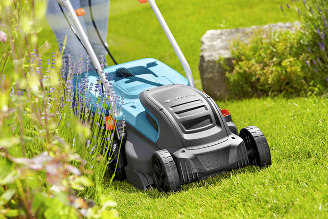 Eine Person bedient einen Rasenmäher in einem Garten neben blühenden Pflanzen.