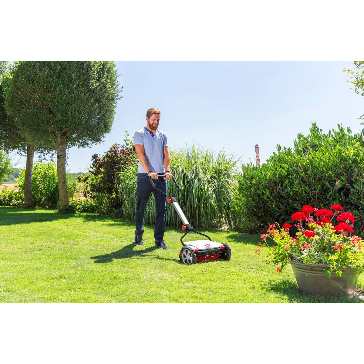 Ein Mann mäht den Rasen mit einem Handrasenmäher im Garten bei Sonnenschein.