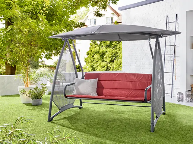 Eine rote Hollywoodschaukel steht mit einem grauen Dach auf einer Terrasse im Garten.