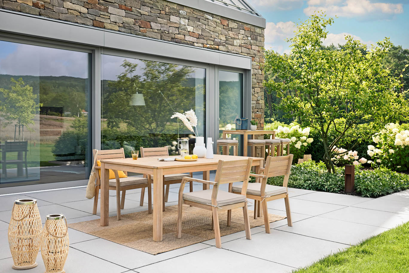 Ein Holztisch mit Stühlen steht auf einer Terrasse neben einem modernen Haus mit Garten.