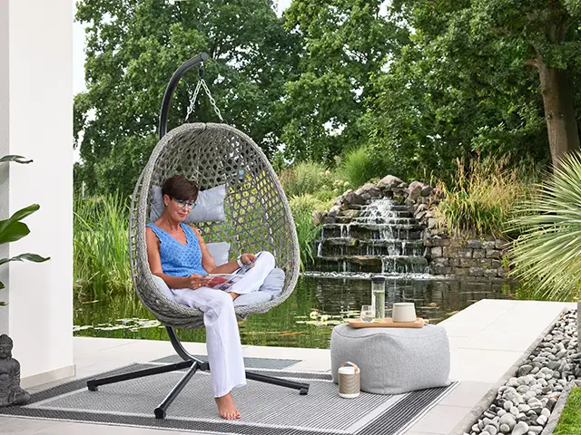 Eine Person sitzt in einem Hängestuhl und liest in einem Garten mit eigenem Teich mit Wasserfall.