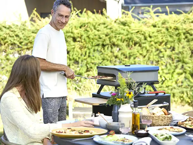 Ein Mann backt Pizza im Freien neben einer Frau an einem Tisch mit Essen.