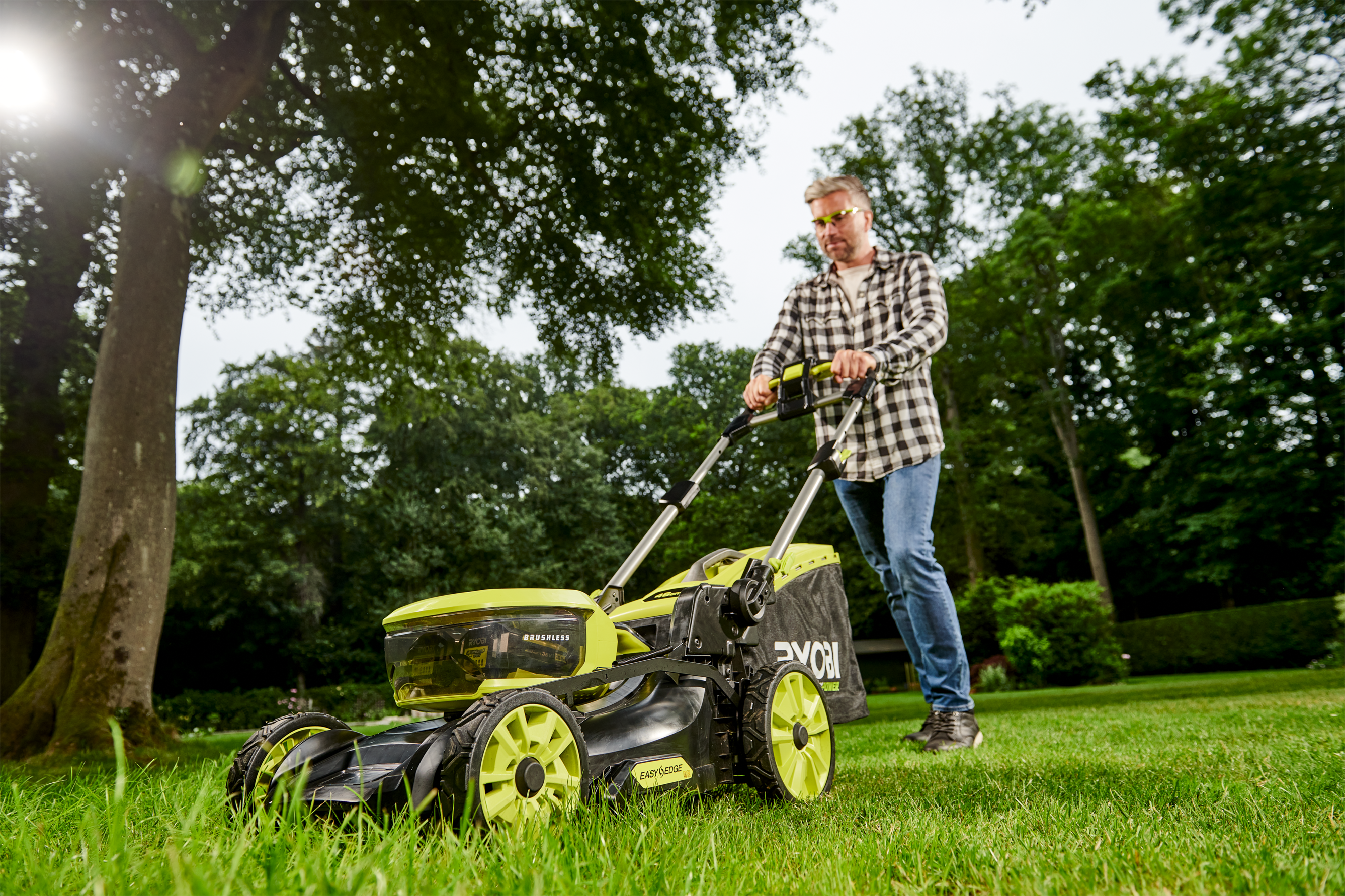 Ein Mann mäht mit einem Rasenmäher auf einer grünen Wiese vor einem Wald.