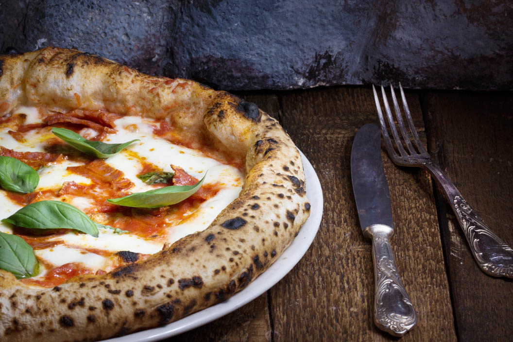 Eine Pizza mit Basilikumblättern liegt neben Besteck auf einem Holztisch.