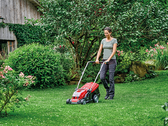 Frau mäht Rasen mit Rasenmäher vor einem Holzhaus