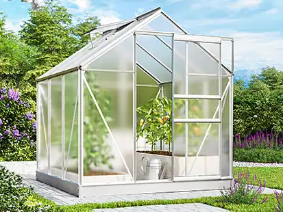 Ein Gewächshaus aus Glas und Metall steht im Garten, umgeben von Pflanzen und Bäumen.