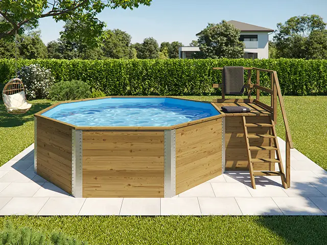 Ein achteckiger Holzpool steht auf Steinplatten in einem grünen Garten mit Bäumen.