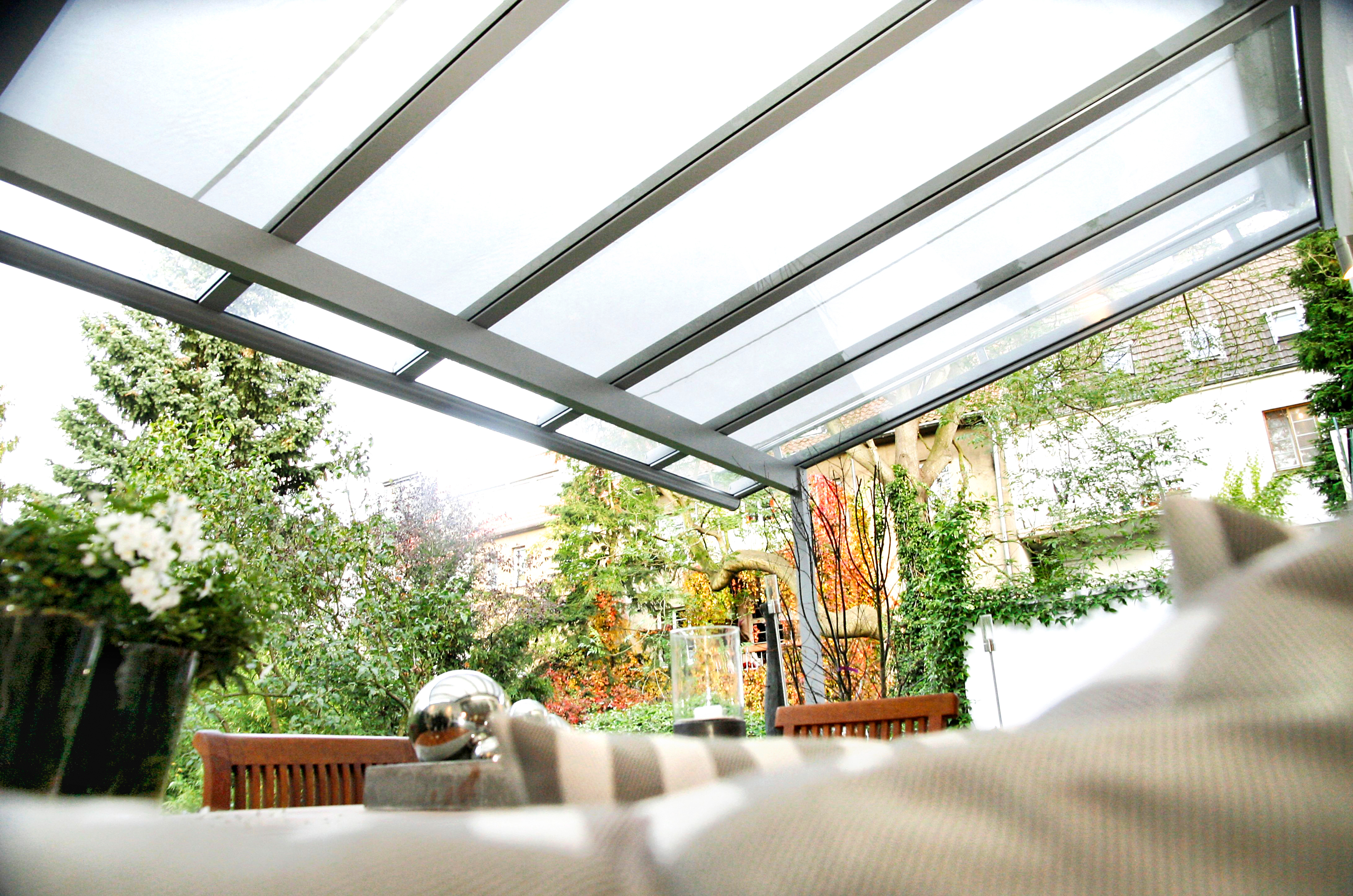 Eine transparente Überdachung schützt eine Terrasse mit Pflanzen und Möbeln in einem grünen, blühenden Garten.